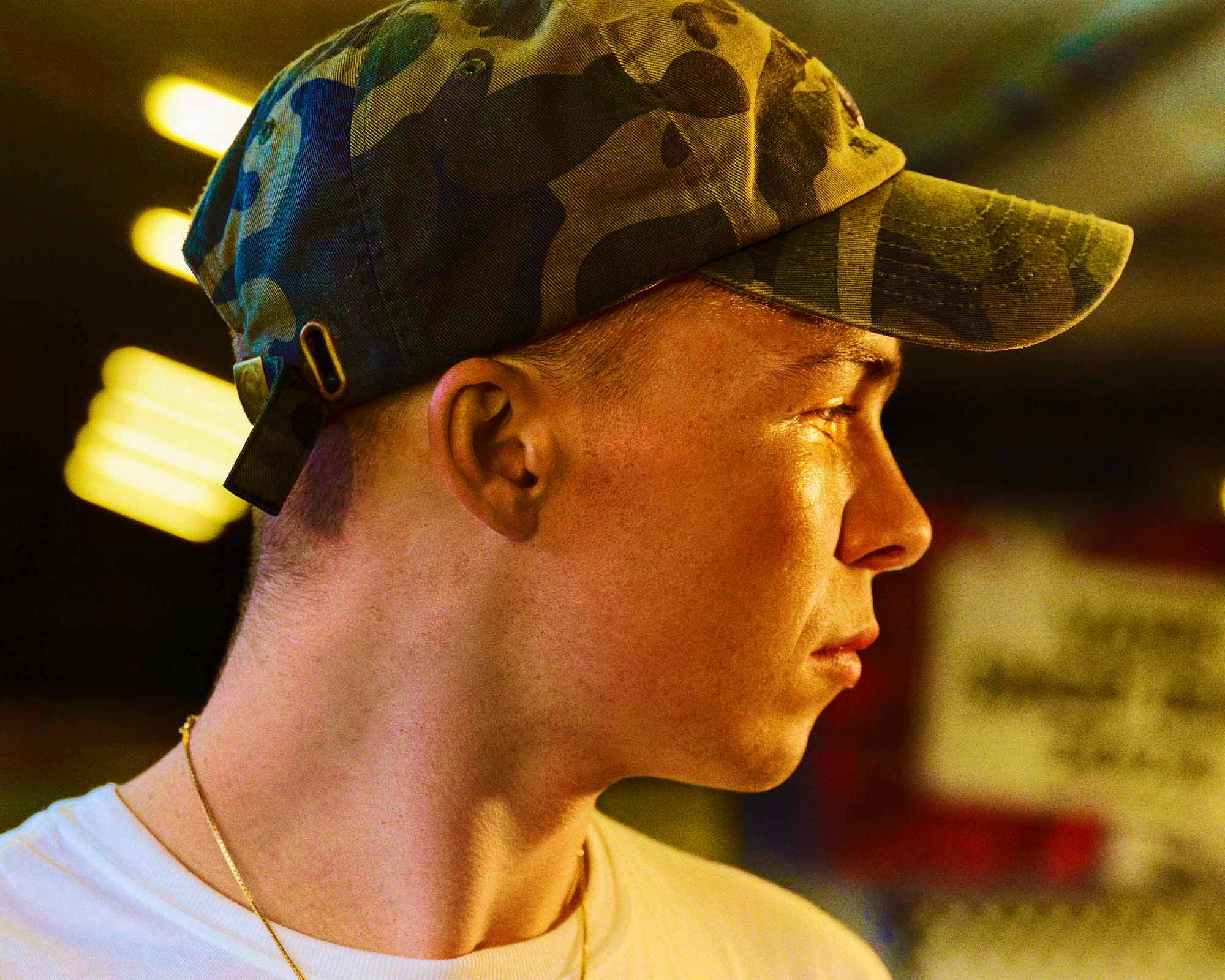 http://Close-up%20profile%20of%20a%20young%20man%20in%20a%20camouflage%20cap%20and%20white%20t-shirt%20with%20a%20gold%20chain,%20looking%20serious,%20against%20a%20blurred%20background%20of%20warm%20lights