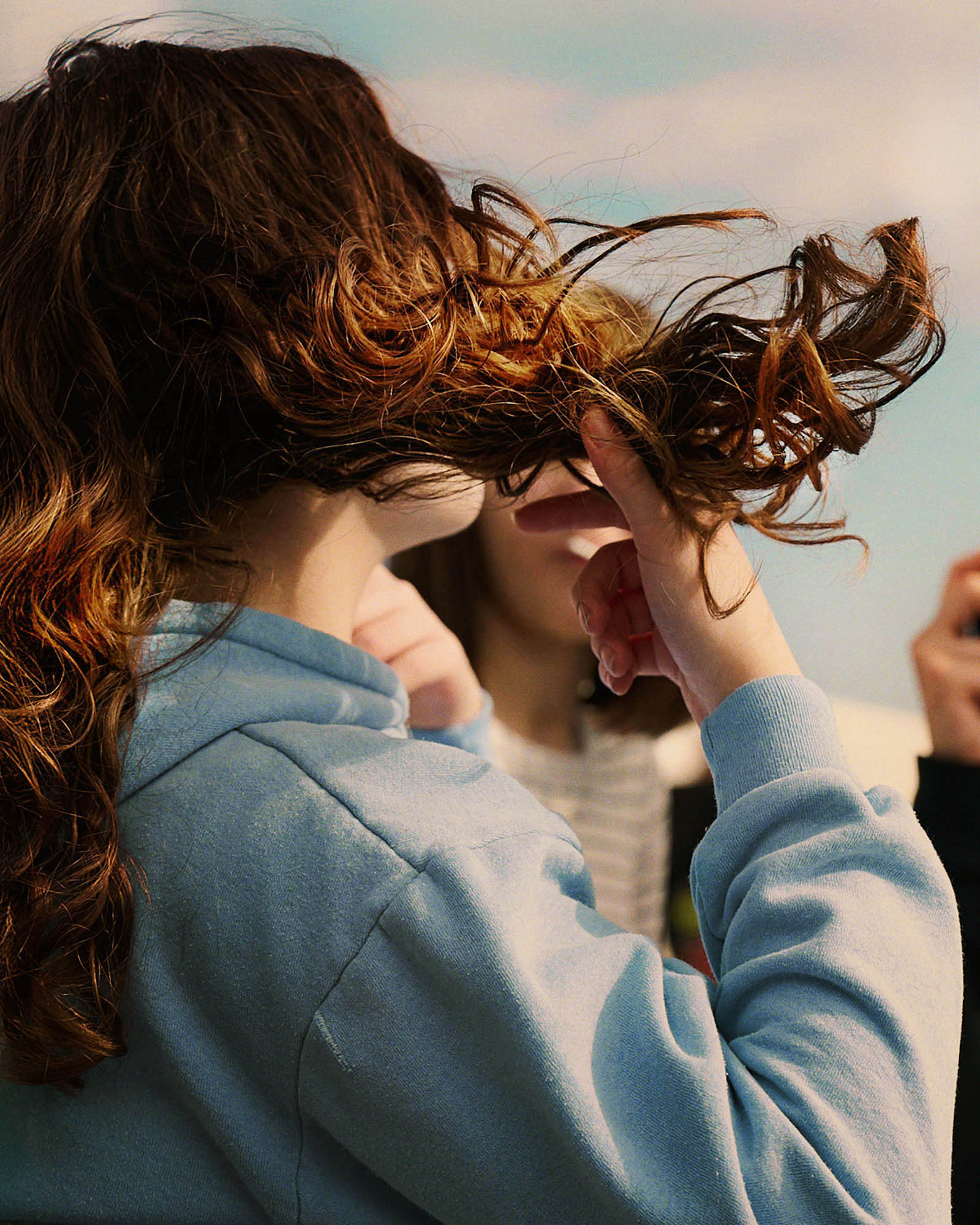 http://A%20young%20woman%20in%20a%20blue%20hoodie,%20seen%20from%20behind,%20flipping%20her%20long%20curly%20brown%20hair,%20with%20a%20blurred%20person%20and%20sky%20in%20the%20background