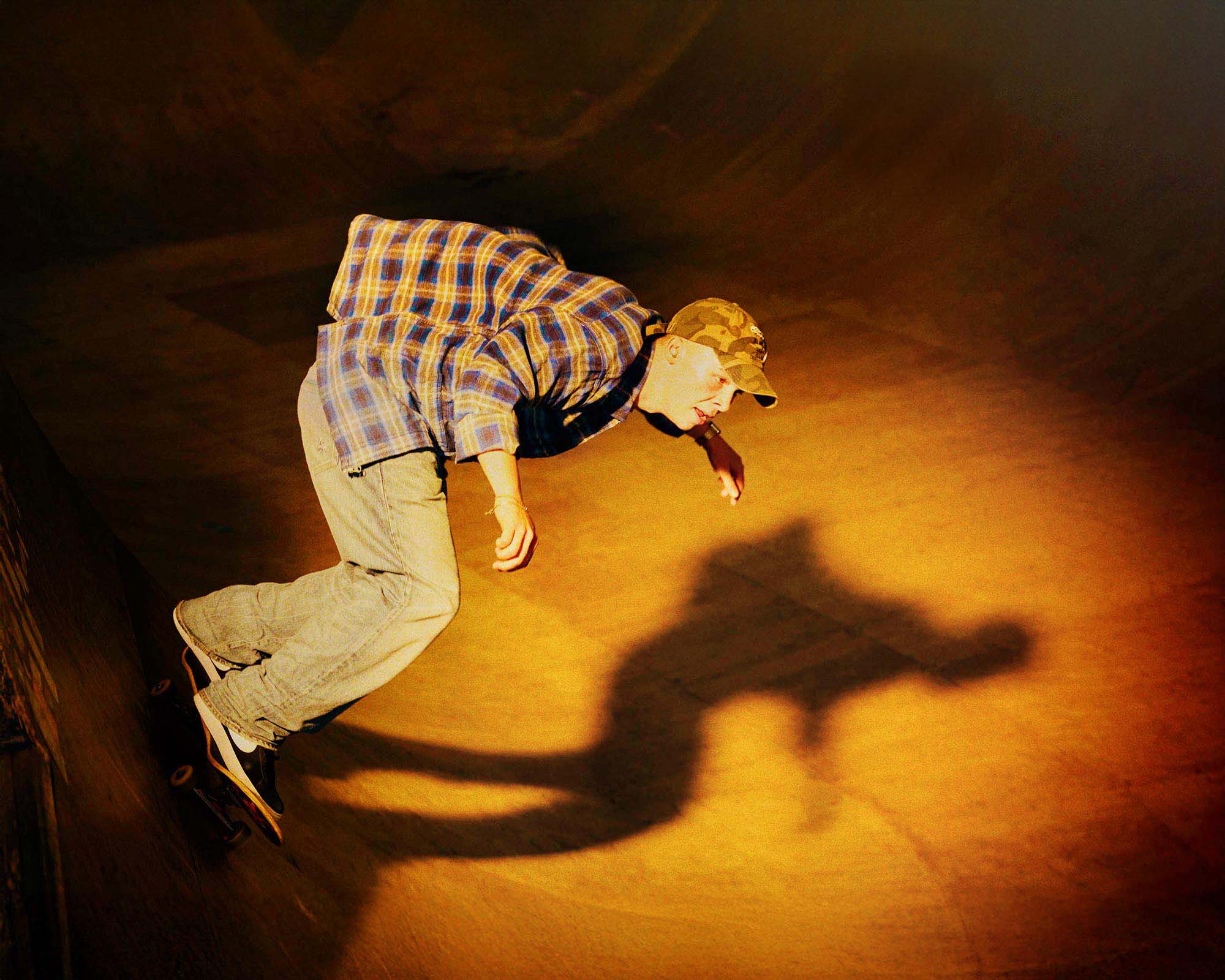 http://Skateboarder%20in%20a%20plaid%20shirt%20and%20camo%20cap%20riding%20in%20a%20wooden%20skate%20bowl,%20with%20a%20dramatic%20shadow%20cast%20by%20overhead%20light.