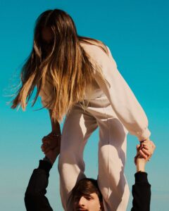 http://A%20low-angle%20shot%20features%20a%20young%20woman%20with%20long,%20light%20brown%20hair,%20wearing%20a%20light-colored%20sweatsuit,%20sitting%20on%20the%20shoulders%20of%20a%20young%20man.%20Her%20face%20is%20obscured%20by%20her%20hair.%20She%20holds%20his%20hands%20for%20balance,%20and%20his%20face%20is%20partially%20visible%20at%20the%20bottom%20of%20the%20frame.%20The%20background%20is%20a%20vibrant,%20clear%20turquoise%20sky,%20suggesting%20a%20sunny%20day.