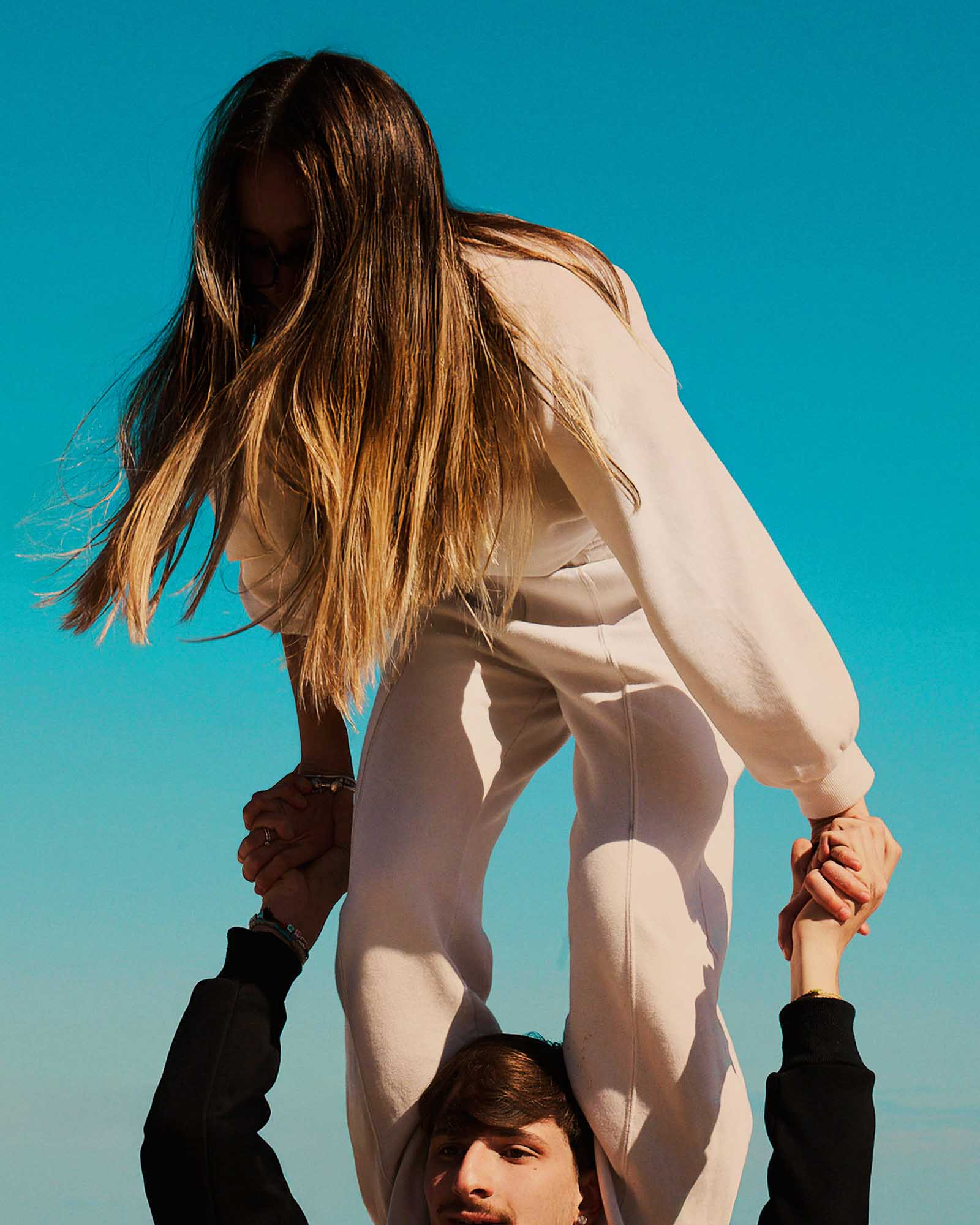 http://A%20low-angle%20shot%20features%20a%20young%20woman%20with%20long,%20light%20brown%20hair,%20wearing%20a%20light-colored%20sweatsuit,%20sitting%20on%20the%20shoulders%20of%20a%20young%20man.%20Her%20face%20is%20obscured%20by%20her%20hair.%20She%20holds%20his%20hands%20for%20balance,%20and%20his%20face%20is%20partially%20visible%20at%20the%20bottom%20of%20the%20frame.%20The%20background%20is%20a%20vibrant,%20clear%20turquoise%20sky,%20suggesting%20a%20sunny%20day.