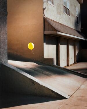 Bright yellow balloon floating in a sunlit urban alleyway against a beige stucco wall, with a concrete ramp in the foreground.